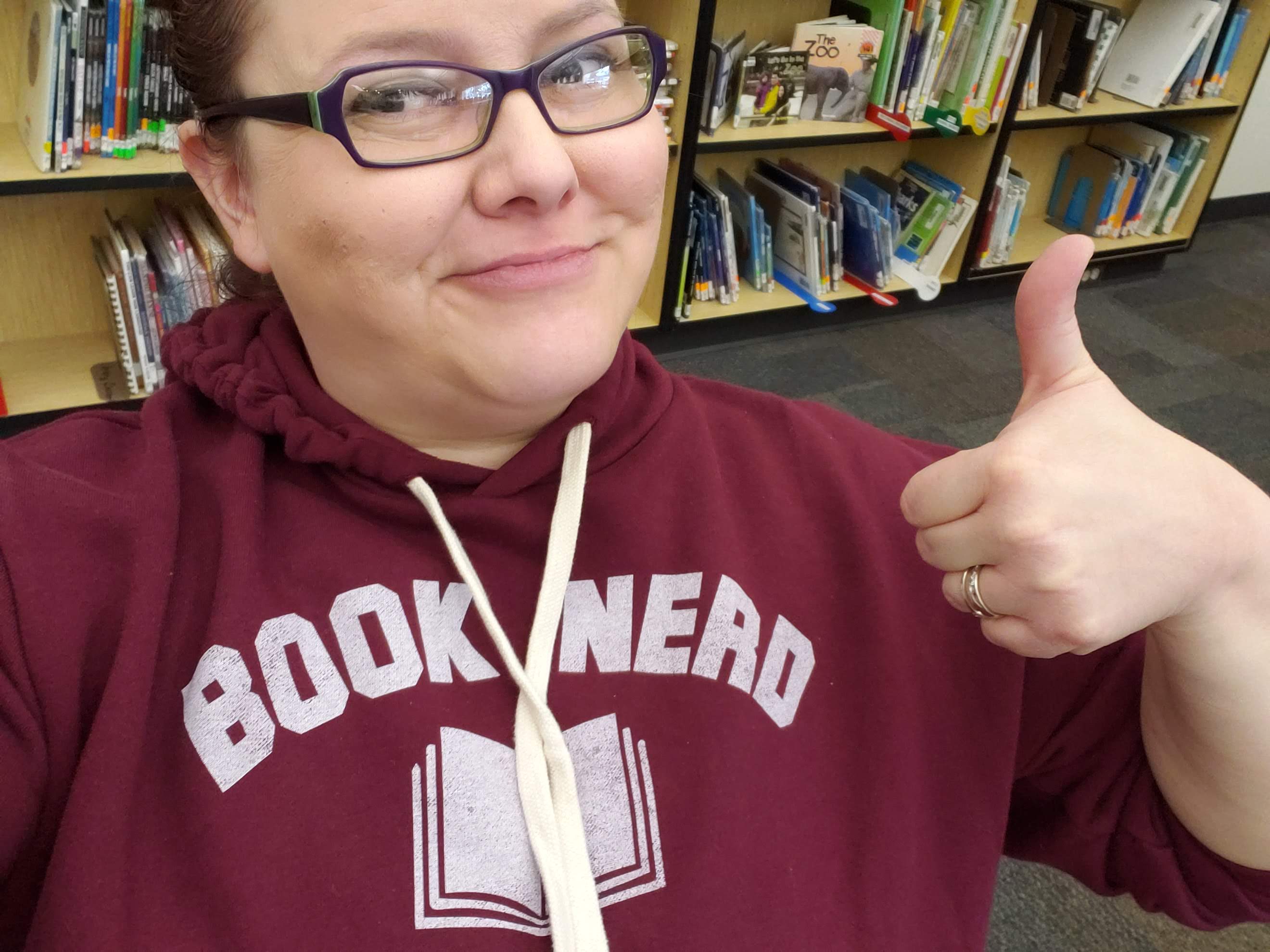 Mrs. Sand wearing a Book Nerd sweatshirt in front of bookshelves with her thumb up 